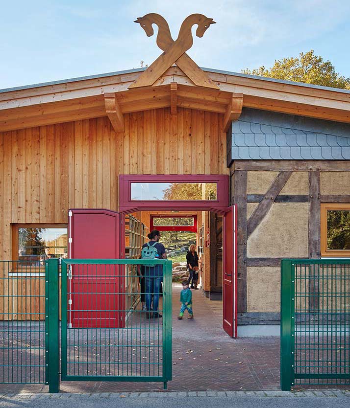 Der Eingang des Stallgebäudes mit roter Tür und Holz-Pferdeköpfen am Giebel.