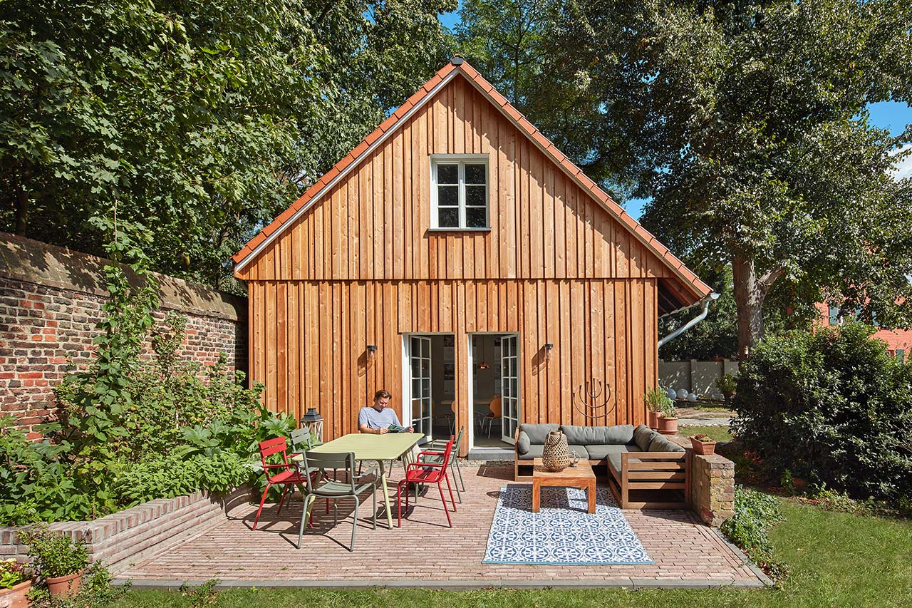 Seitliche Ansicht des Ateliers mit gepflasterter Terrasse und Holzfassade, daneben die alte Backsteinmauer.