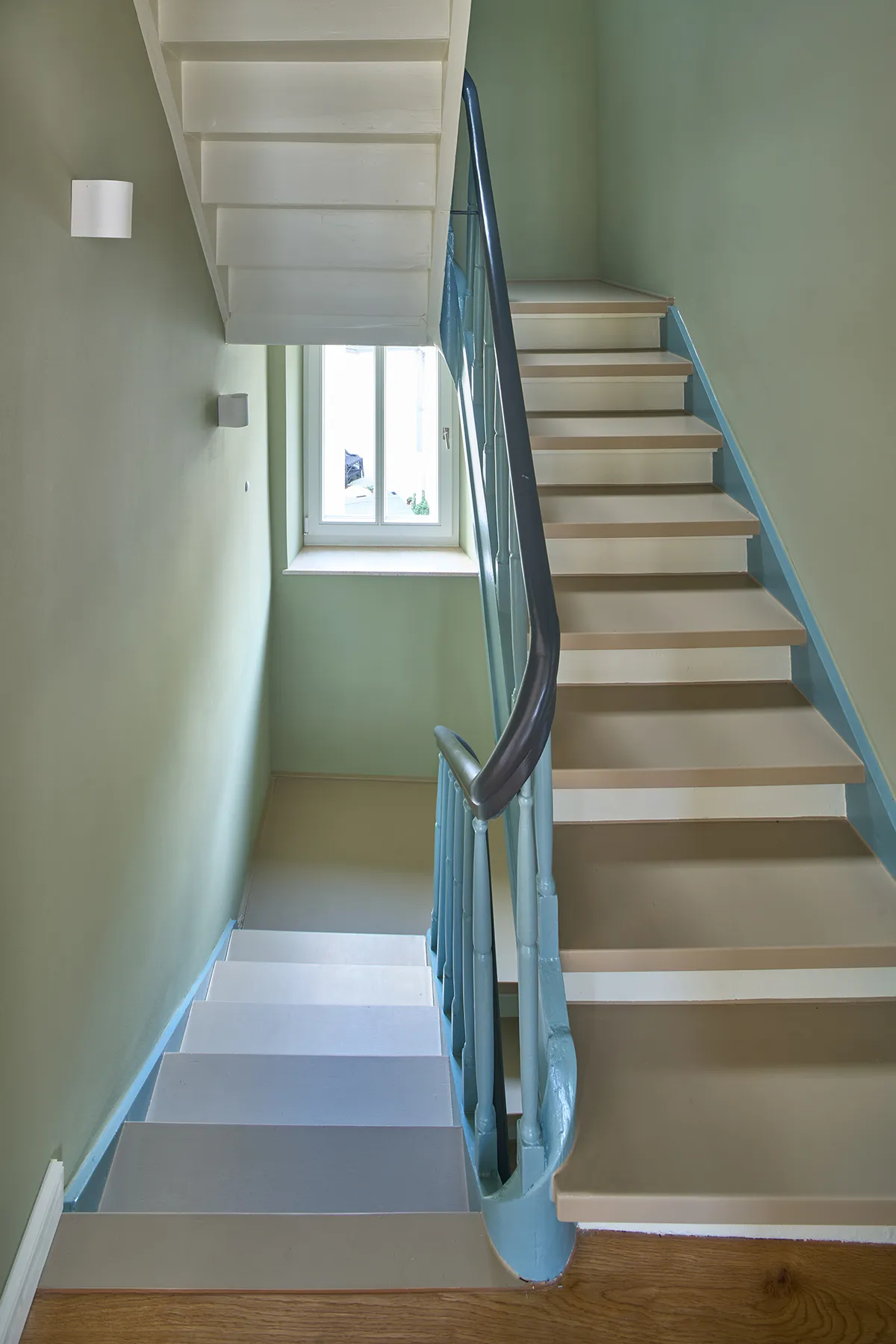 Sanierter Treppenraum mit hellen Wandflächen, erneuerten Stufen und farbig gefasstem Handlauf, belichtet durch Fenster im Treppenhaus.